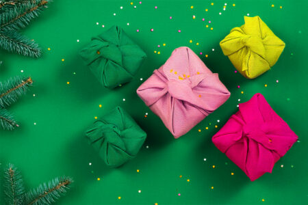 5 cadeaux emballés dans des furoshikis sur un fond vert, saupoudrés de confettis multicolores, avec des branches de sapin sur le côté gauche de l'image